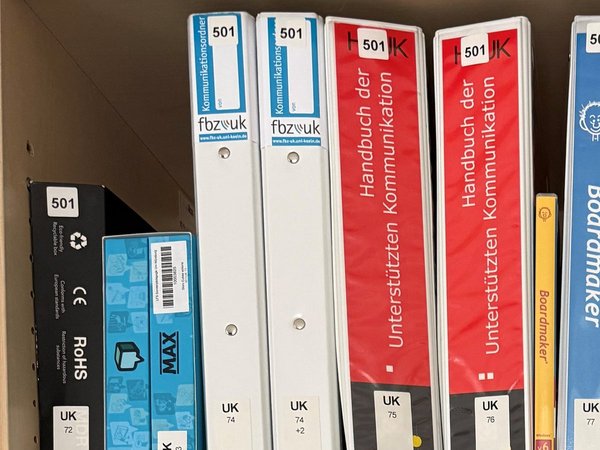 Regal der LFS mit Ordnern und Materialboxen zur Unterstützten Kommunikation, jeweils beschriftet mit Signaturen wie UK 74 und UK 74 +2